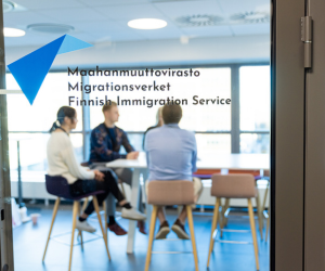 People who are having a conversation. There is a door in the front of the picture that has the Finnish Migration Service's logo on it.